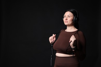 Photo of Plus size woman in gym clothes with headphones and skipping rope on black background, space for text