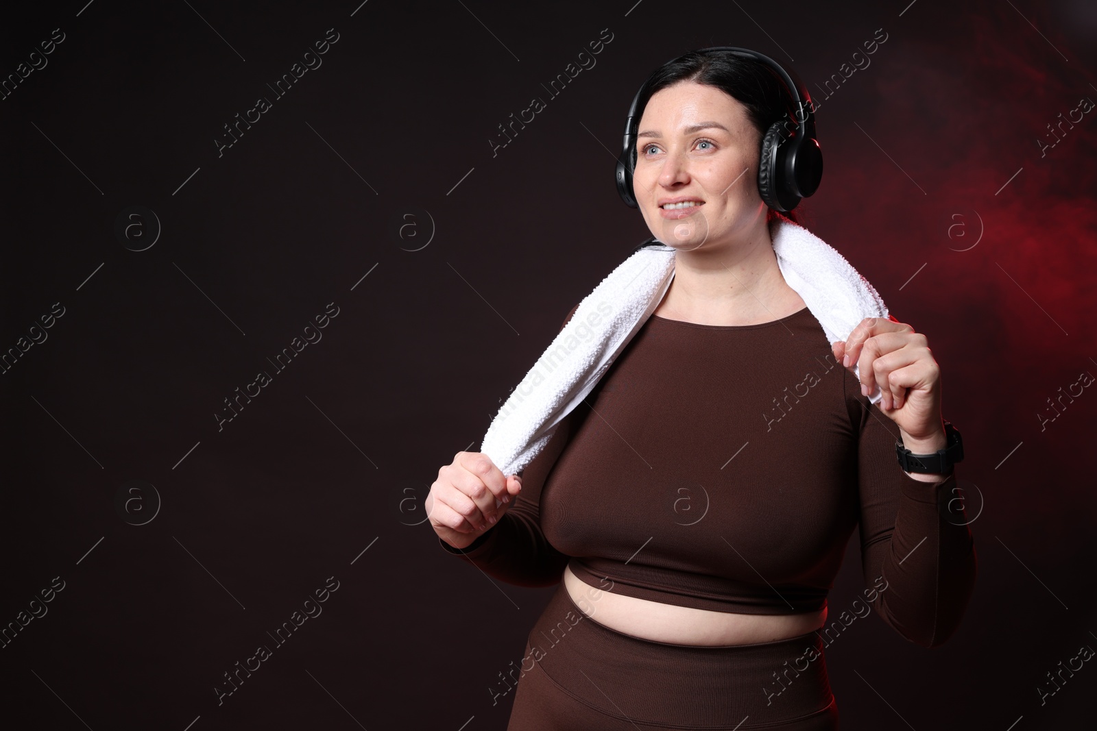 Photo of Plus size woman in gym clothes with headphones and towel on black background with smoke, space for text