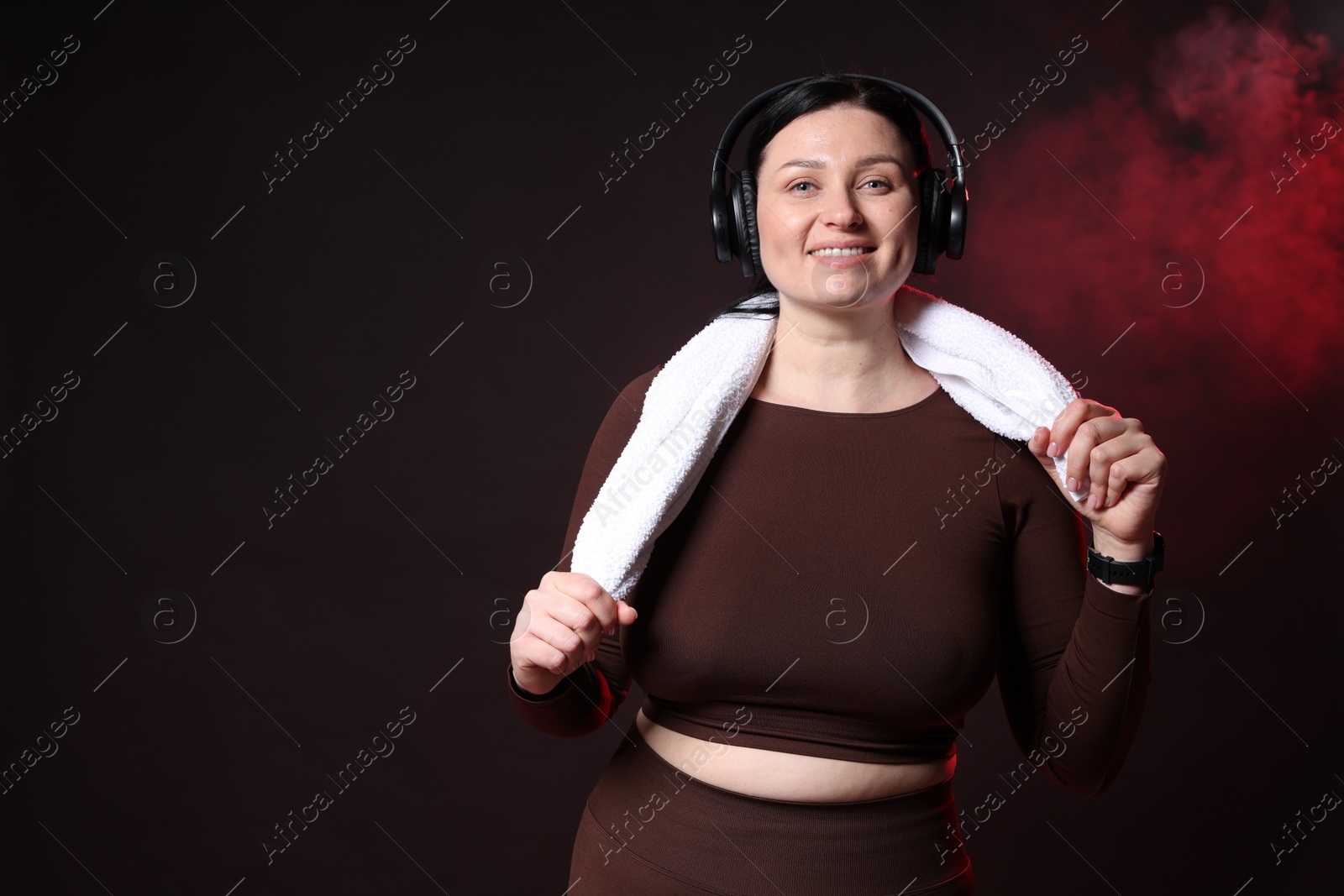 Photo of Plus size woman in gym clothes with headphones and towel on black background with smoke, space for text