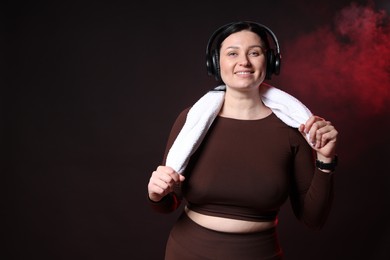 Photo of Plus size woman in gym clothes with headphones and towel on black background with smoke, space for text