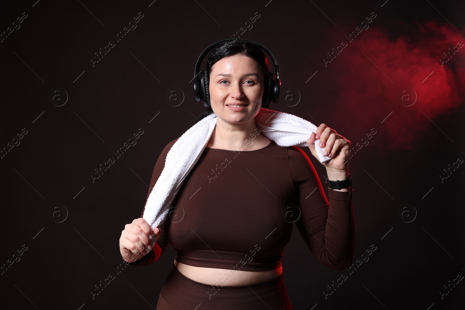 Photo of Plus size woman in gym clothes with headphones and towel on black background with smoke