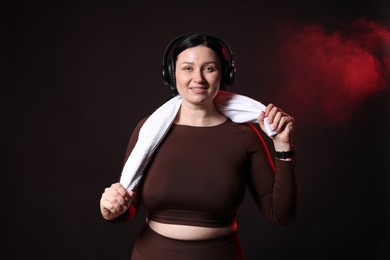 Photo of Plus size woman in gym clothes with headphones and towel on black background with smoke