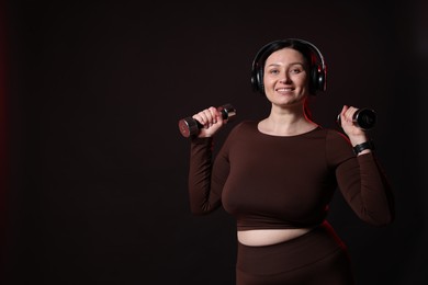 Photo of Plus size woman in gym clothes with dumbbells and headphones on black background, space for text