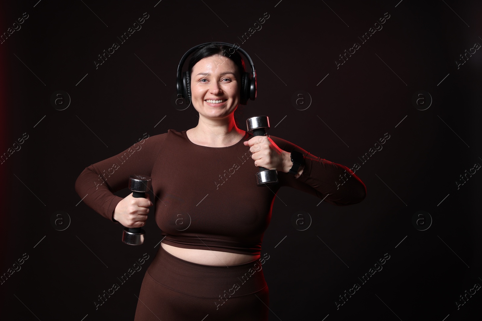 Photo of Plus size woman in gym clothes with dumbbells and headphones on black background