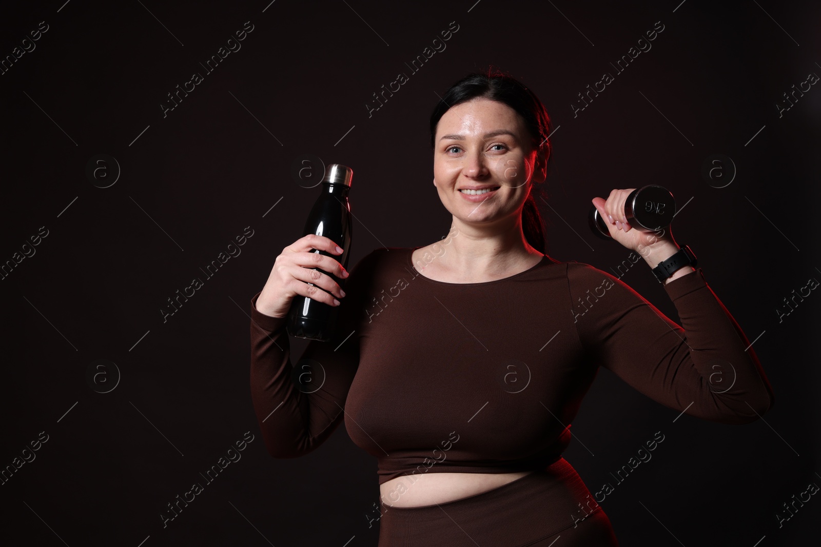 Photo of Plus size woman in gym clothes with dumbbell and water bottle on black background, space for text