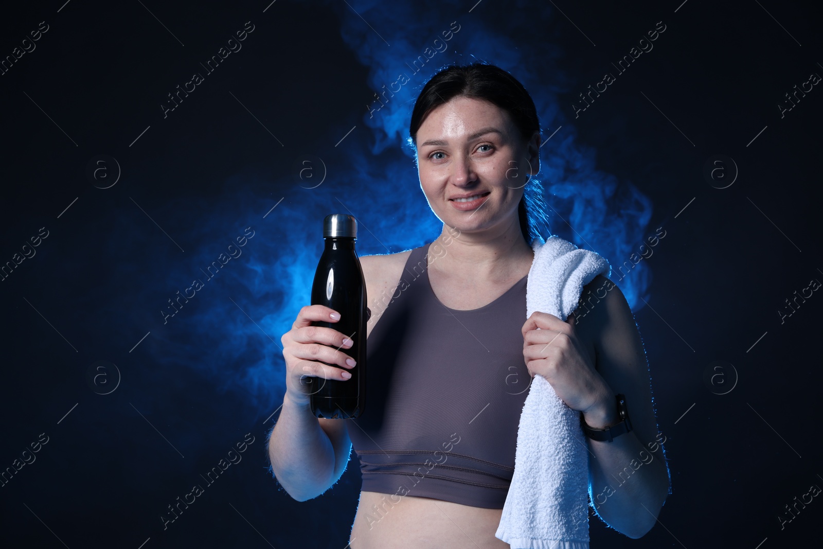 Photo of Plus size woman in gym clothes with water bottle and towel on dark blue background with smoke