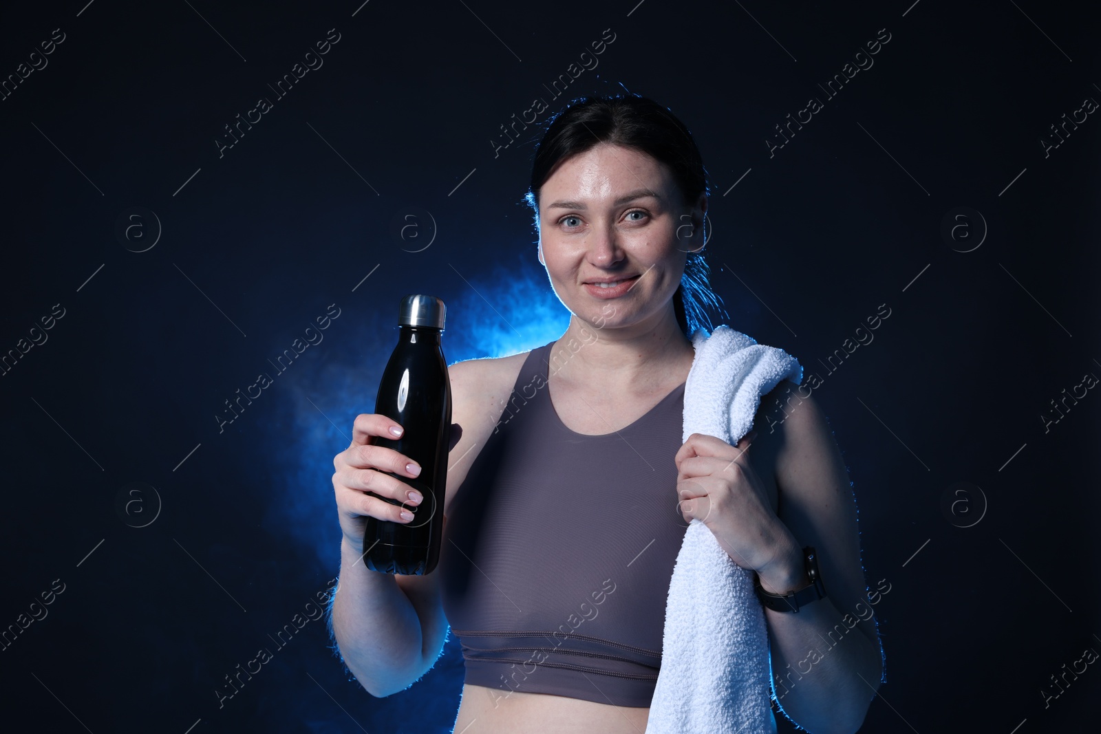 Photo of Plus size woman in gym clothes with water bottle and towel on dark blue background with smoke