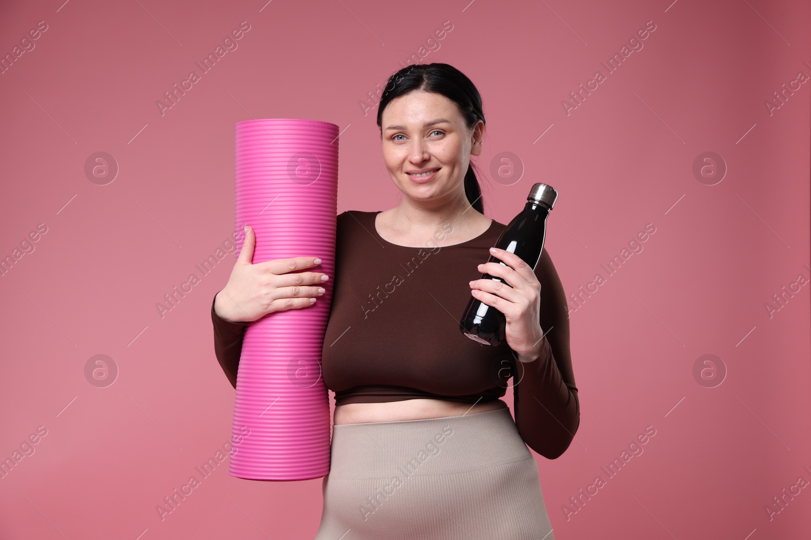 Photo of Plus size woman in gym clothes with fitness mat and water bottle on pink background