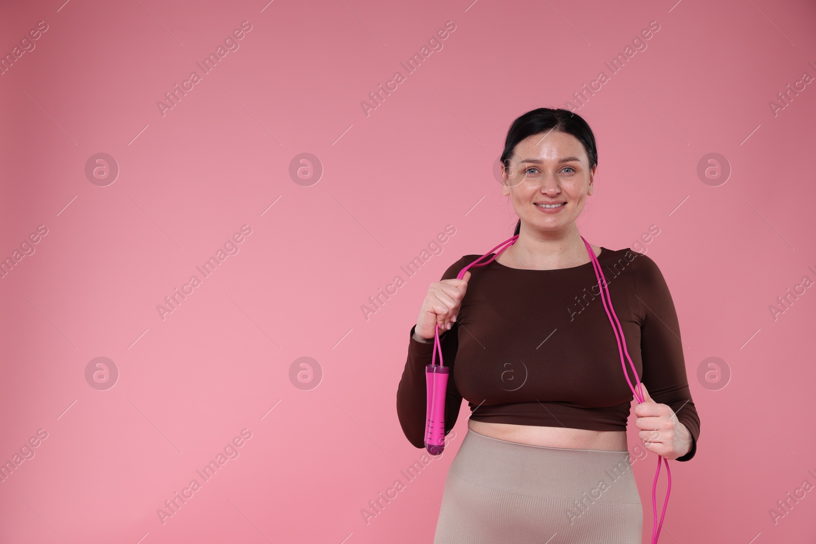 Photo of Plus size woman in gym clothes with skipping rope on pink background, space for text