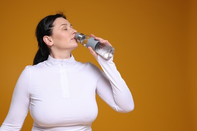 Photo of Plus size woman in gym clothes drinking water on orange background, space for text