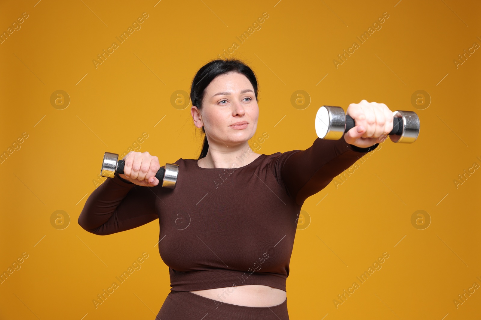 Photo of Plus size woman in gym clothes with dumbbells on orange background