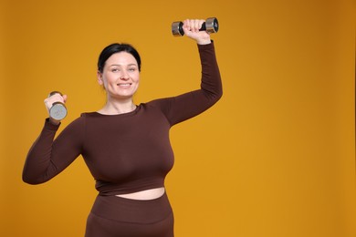 Photo of Plus size woman in gym clothes with dumbbells on orange background, space for text