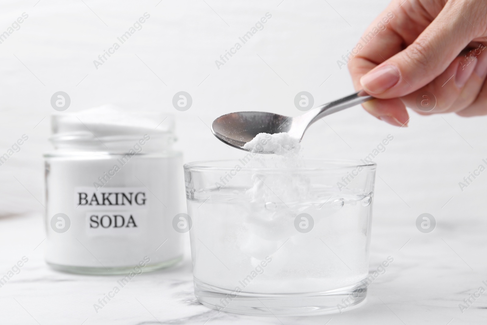 Photo of Woman adding baking soda to glass of water at white marble table, closeup