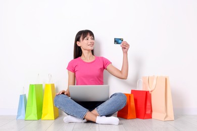 Internet shopping. Happy woman with credit card, laptop and colorful bags sitting on floor near white wall