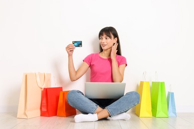 Photo of Internet shopping. Happy woman with credit card, laptop and colorful bags sitting on floor near white wall