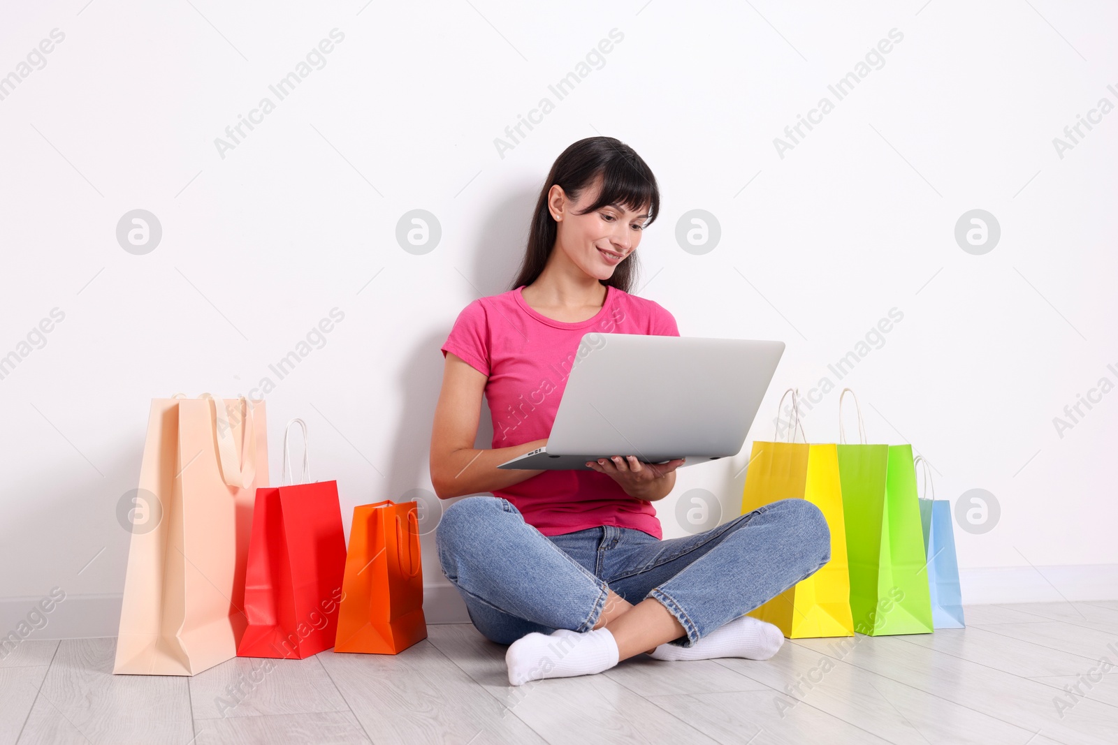 Photo of Internet shopping. Happy woman with laptop and colorful bags sitting on floor near white wall