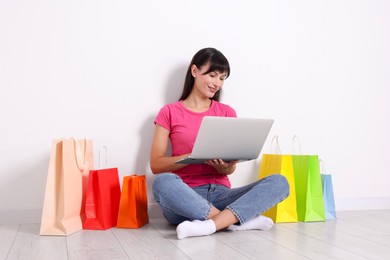 Internet shopping. Happy woman with laptop and colorful bags sitting on floor near white wall