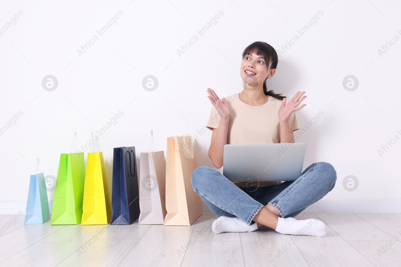 Photo of Internet shopping. Happy woman with laptop and colorful bags sitting on floor near white wall