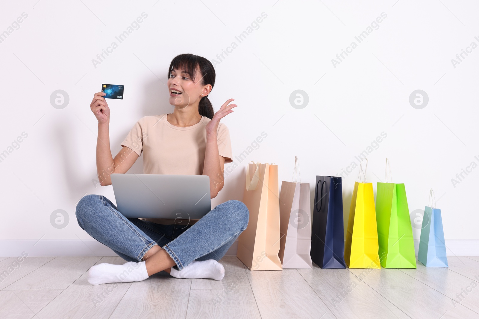 Photo of Internet shopping. Happy woman with laptop and colorful bags sitting on floor near white wall