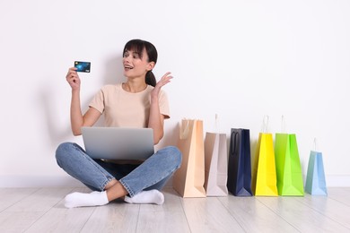 Internet shopping. Happy woman with laptop and colorful bags sitting on floor near white wall