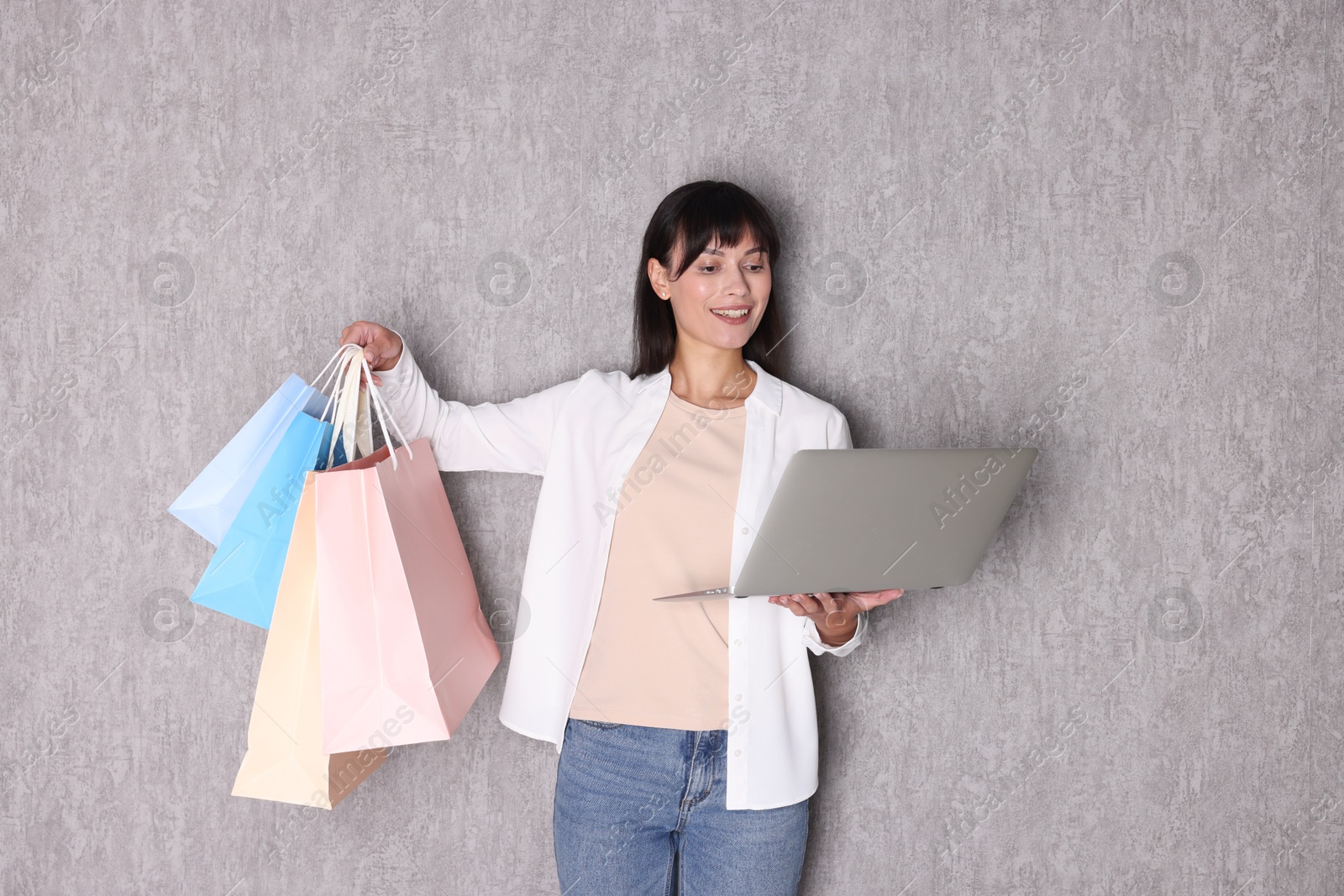 Photo of Internet shopping. Happy woman with laptop and colorful bags near grey wall