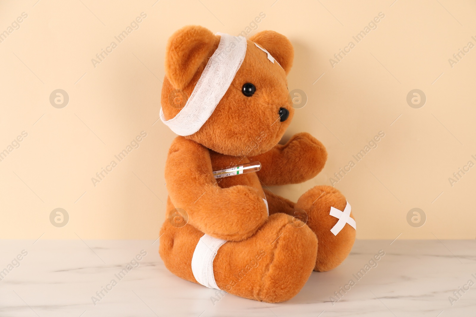 Photo of Teddy bear with bandages, sticking plasters and thermometer on white marble table against beige background