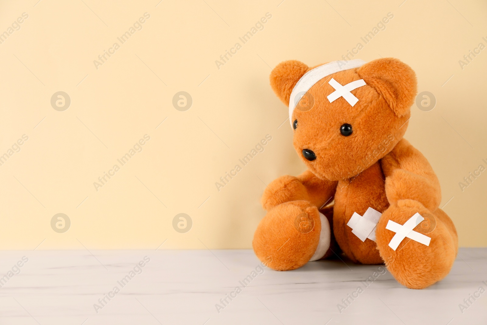 Photo of Teddy bear with bandages and sticking plasters on white marble table against beige background, space for text