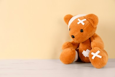 Photo of Teddy bear with bandages and sticking plasters on white marble table against beige background, space for text