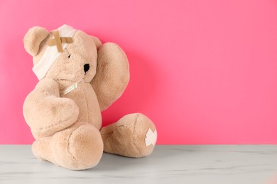 Photo of Teddy bear with bandage, sticking plasters and thermometer on white marble table against pink background, space for text