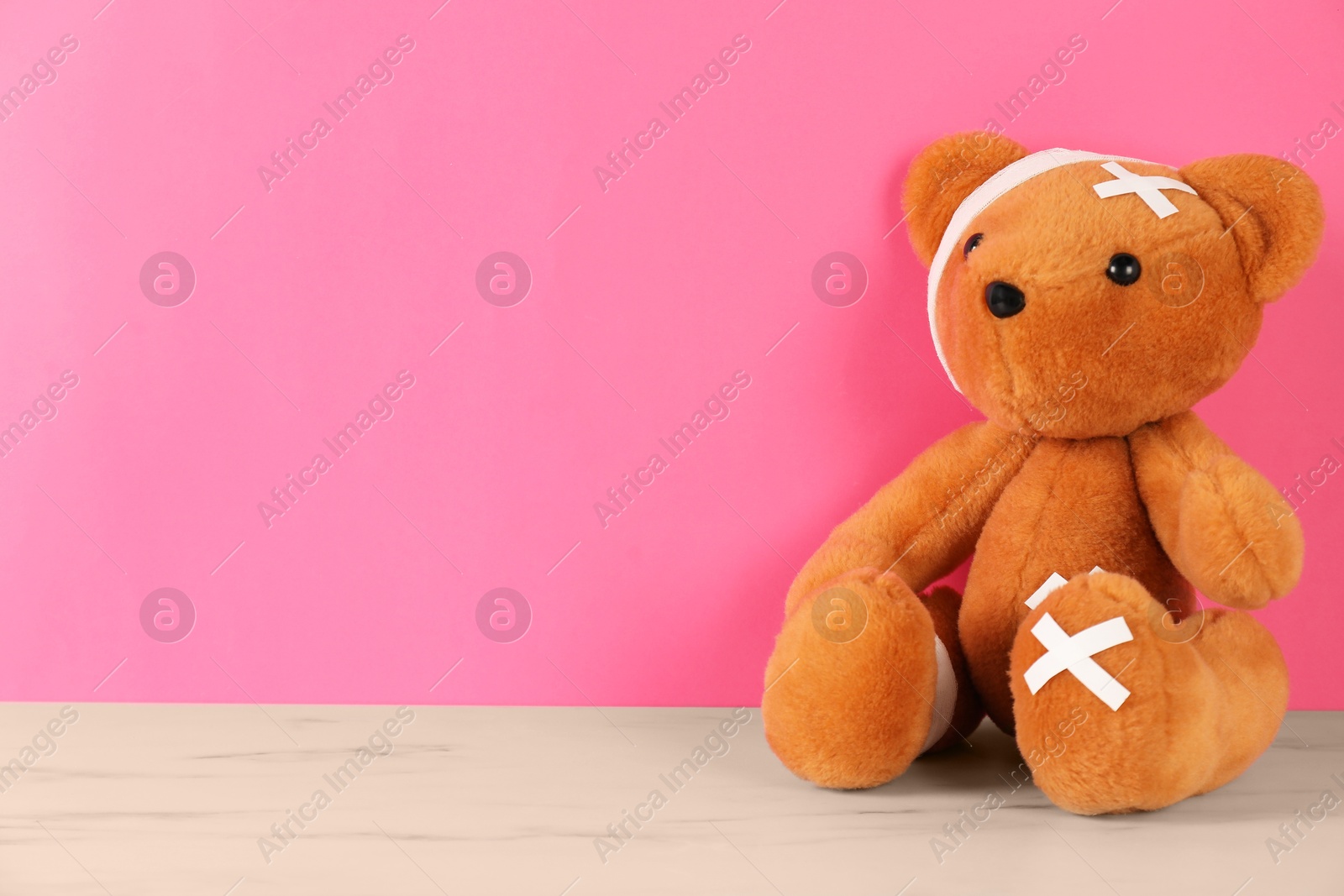 Photo of Teddy bear with bandages and sticking plasters on white marble table against pink background, space for text