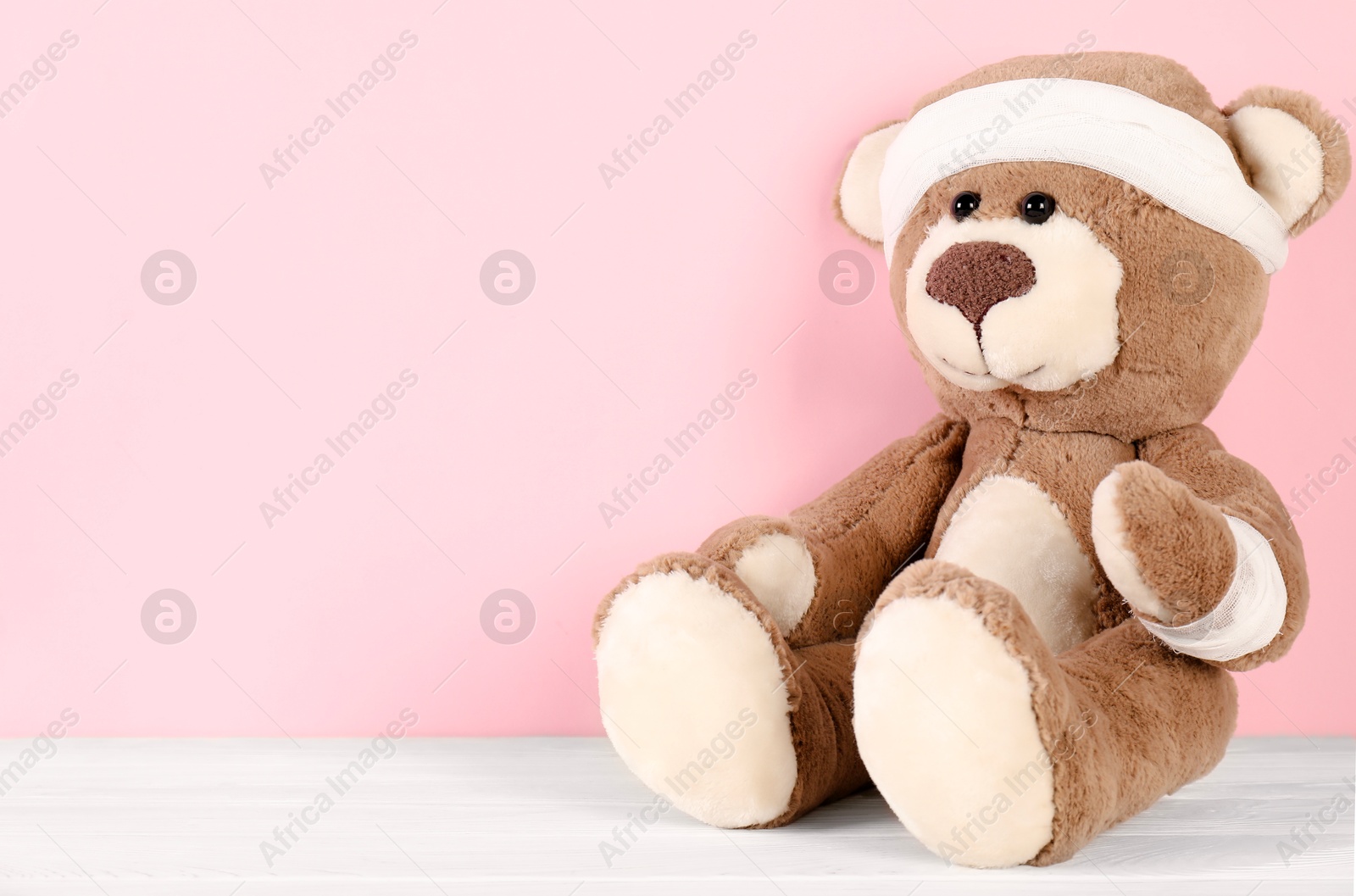 Photo of Teddy bear with bandages on white wooden table against pink background, space for text