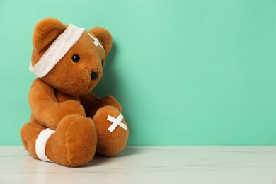 Photo of Teddy bear with bandages and sticking plasters on white marble table against turquoise background, space for text
