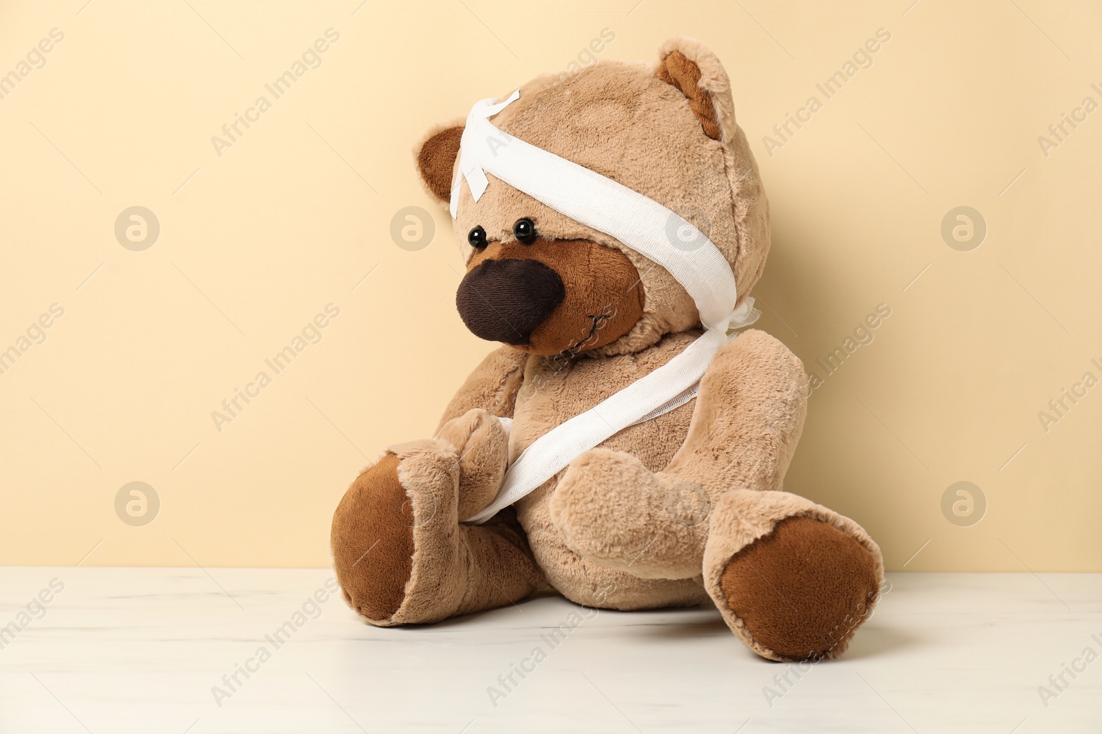Photo of Teddy bear with bandages and sticking plasters on white marble table against beige background