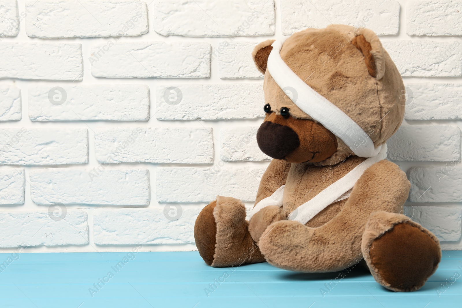 Photo of Teddy bear with bandages on light blue wooden table against white brick wall, space for text