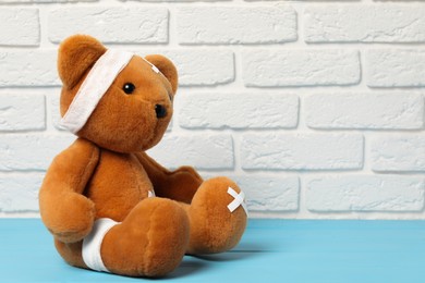 Photo of Teddy bear with bandages and sticking plasters on light blue wooden table against white brick wall, space for text