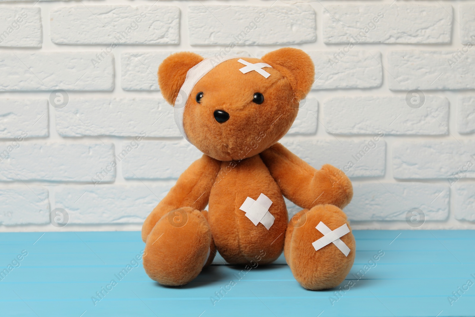 Photo of Teddy bear with bandages and sticking plasters on light blue wooden table against white brick wall