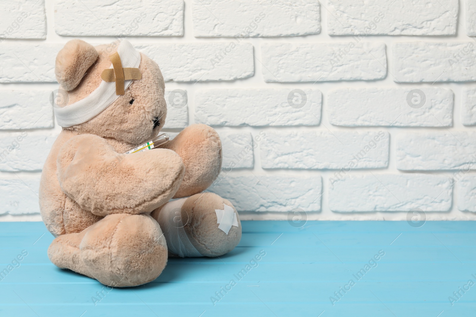 Photo of Teddy bear with bandages, thermometer and sticking plasters on light blue wooden table against white brick wall, space for text