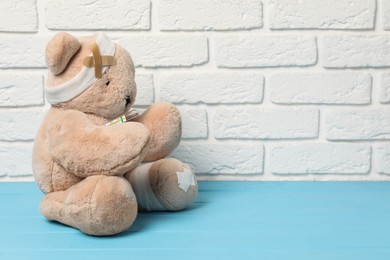 Photo of Teddy bear with bandages, thermometer and sticking plasters on light blue wooden table against white brick wall, space for text
