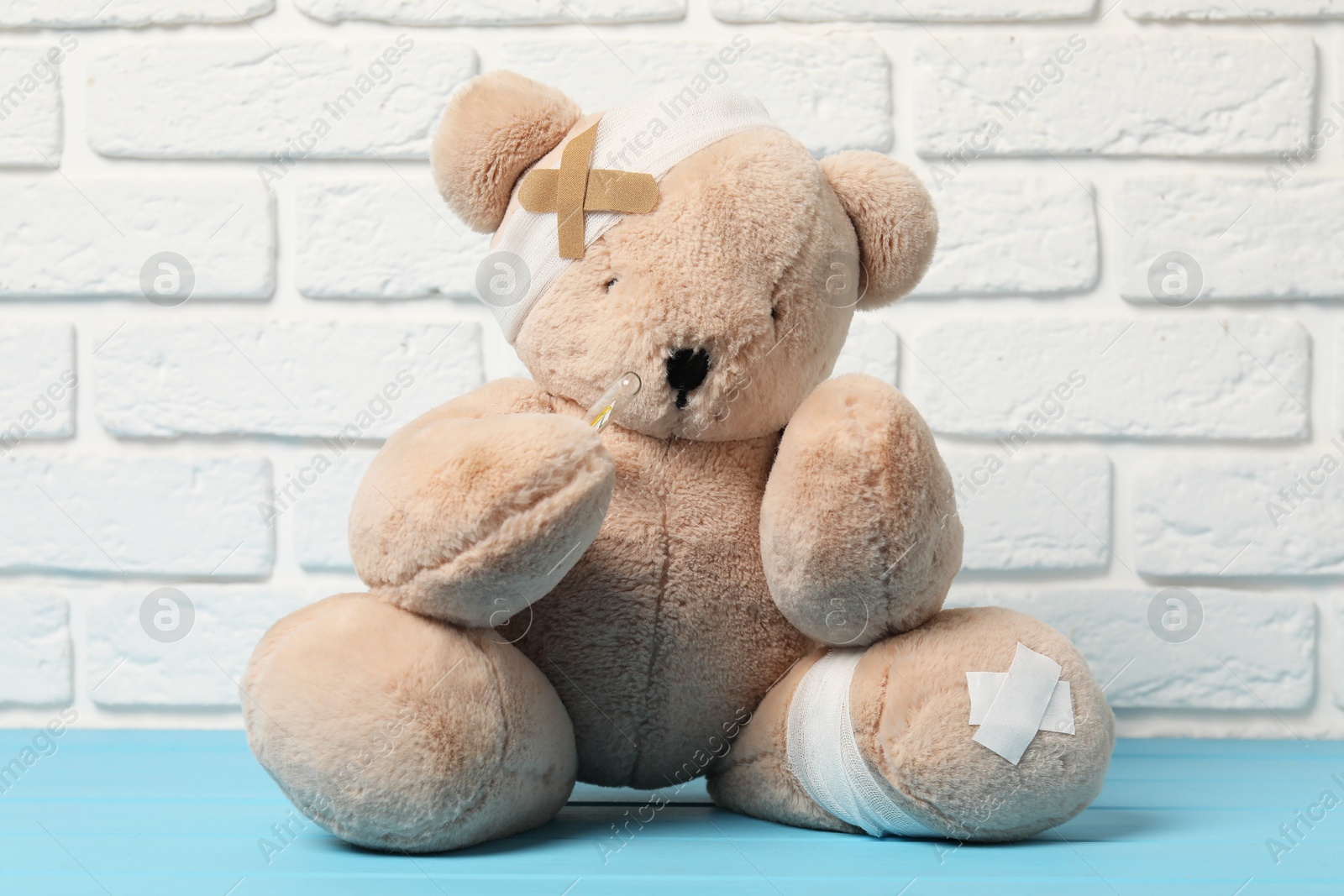 Teddy bear with bandages, thermometer and sticking plasters on light blue wooden table against white brick wall Photo of Teddy bear with bandages, thermometer and sticking plasters on light blue wooden table against white brick wall