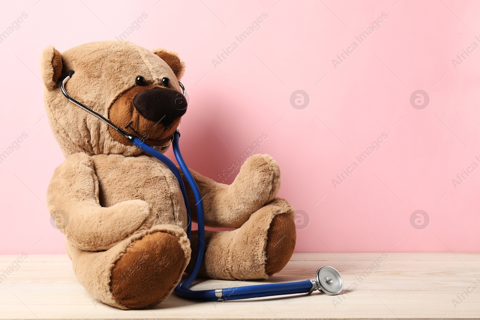 Photo of Pediatrics concept. Teddy bear with stethoscope on wooden table against pink background, space for text