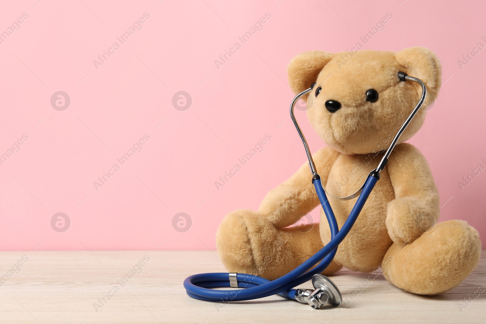 Photo of Pediatrics concept. Teddy bear with stethoscope on wooden table against pink background, space for text