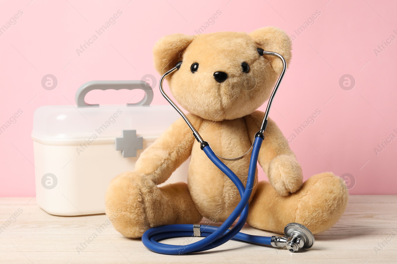 Photo of Pediatrics concept. Teddy bear with stethoscope and first aid kit on wooden table against pink background