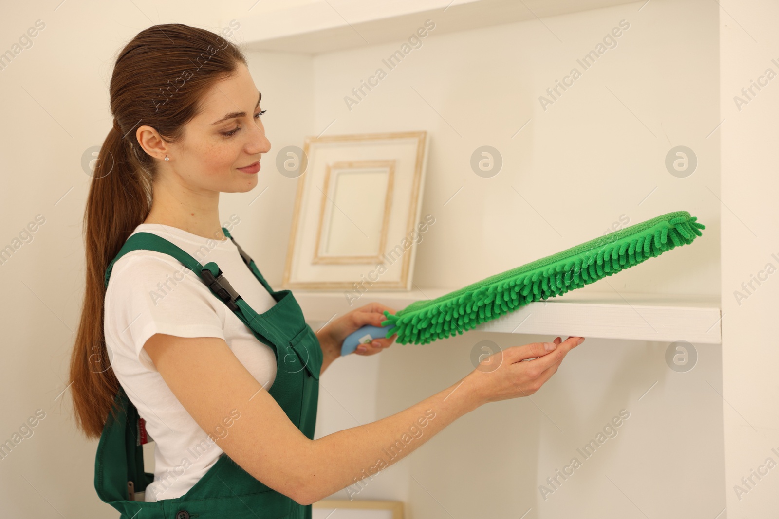 Photo of Professional janitor cleaning dust off shelves indoors