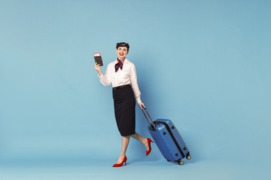 Happy stewardess with suitcase, passport and flight tickets on light blue background