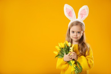 Photo of Cute little girl with bunny ears and tulips on orange background, space for text. Easter celebration