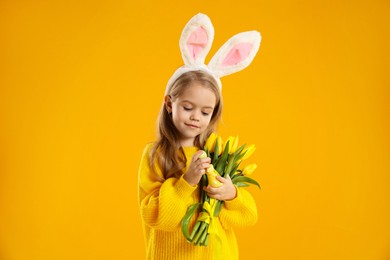 Photo of Cute little girl with bunny ears and tulips on orange background. Easter celebration