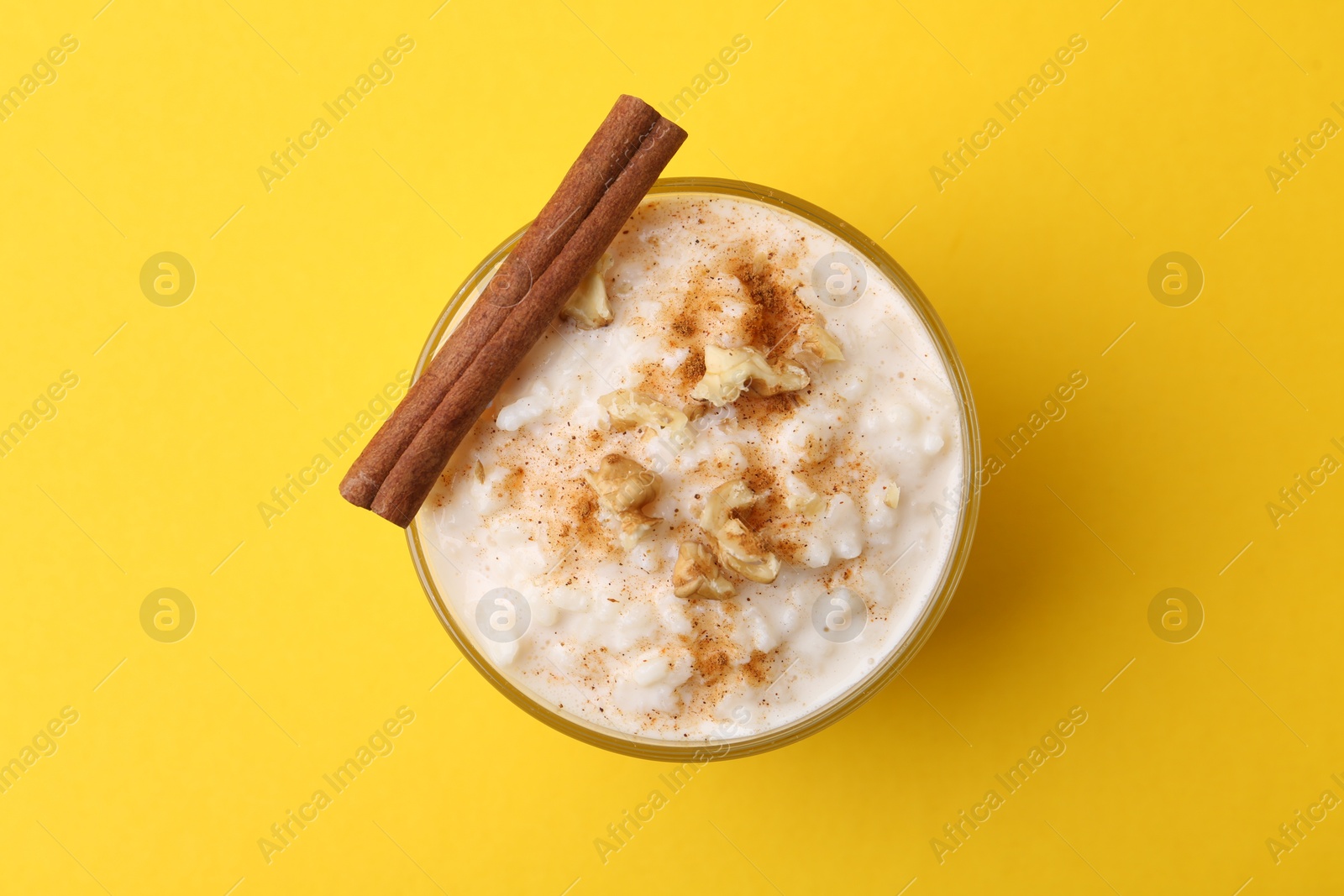 Photo of Tasty rice pudding with cinnamon and nuts in glass bowl on yellow background, top view