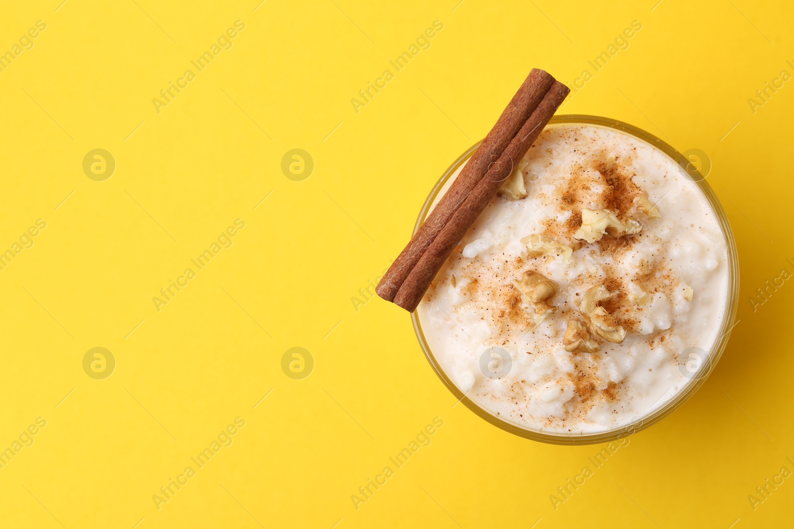 Photo of Tasty rice pudding with cinnamon and nuts in glass bowl on yellow background, top view. Space for text