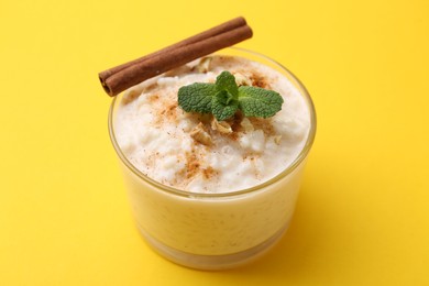 Photo of Tasty rice pudding with cinnamon, mint and nuts in glass bowl on yellow background, closeup