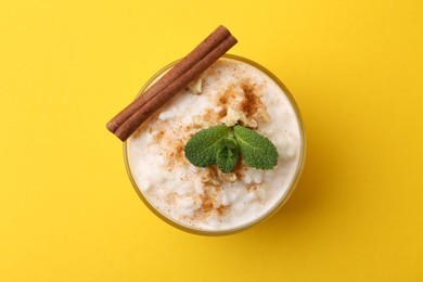 Photo of Tasty rice pudding with cinnamon, mint and nuts in glass bowl on yellow background, top view